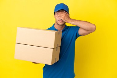 Delivery man over isolated yellow wall covering eyes by hands. Do not want to see something