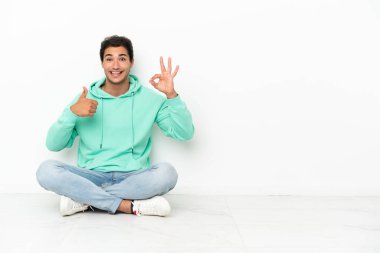 Beyaz, yakışıklı bir adam yerde oturmuş işaret veriyor ve el hareketi yapıyor.