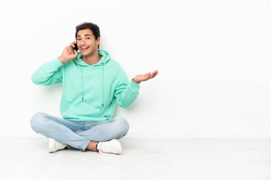 Beyaz tenli yakışıklı adam yerde oturuyor ve cep telefonuyla biriyle sohbet ediyor.