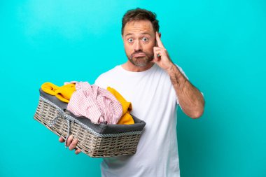 Elinde çamaşır sepeti olan orta yaşlı bir kadın mavi arka planda izole edilmiş bir fikir düşünüyor.