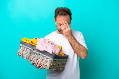 Elinde çamaşır sepeti olan orta yaşlı bir kadın yorgun ve hasta bir ifadeyle mavi arka planda izole edilmiş.