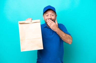 Orta yaşlı bir adam mavi arka planda izole edilmiş bir paket yiyecek alıyor mutlu ve ağzı eliyle kaplayan bir gülümseme.