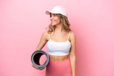 Genç sporcu kadın yoga derslerine giderken elinde minderle yan tarafa bakıyor ve gülümsüyor.