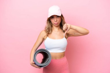 Genç sporcu kadın yoga derslerine giderken elinde paspas tutarken olumsuz bir ifadeyle baş parmağını aşağı gösteriyor.