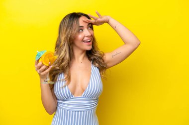 Sarı arka planda mayo giymiş ve elinde kokteyl tutan beyaz bir kadın.