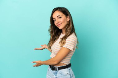 İzole edilmiş mavi arka planda genç bir kadın. Davet etmek için ellerini yana uzatıyor.