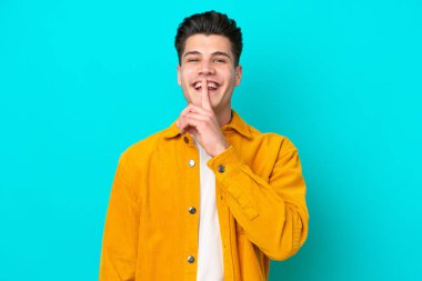 Genç, beyaz, mavi bakkalda izole edilmiş genç bir adam sessizlik işareti gösteriyor. Parmağını ağzına sok.