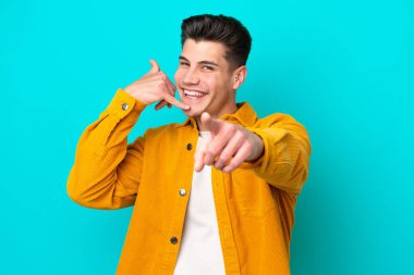 Genç, yakışıklı, beyaz bir adam, mavi bir bakkalda izole edilmiş, telefon jesti yapıyor ve ön tarafı işaret ediyor.