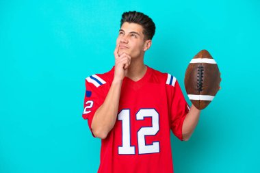 Rugby oynayan Kafkasyalı genç adam mavi arka planda izole edilmiş, yukarı bakarken şüpheleri var.