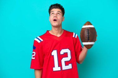 Rugby oynayan genç beyaz adam mavi arka planda izole edilmiş yukarı bakıyor ve şaşırmış bir ifadeyle