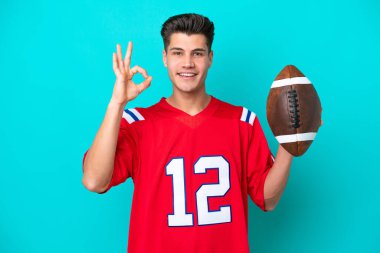 Genç beyaz adam rugby oynuyor. Mavi arka planda izole edilmiş. Parmaklarıyla işaret veriyor.