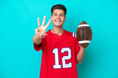 Mavi arka planda ragbi oynayan genç beyaz adam mutlu ve parmaklarıyla üçe kadar sayıyor.