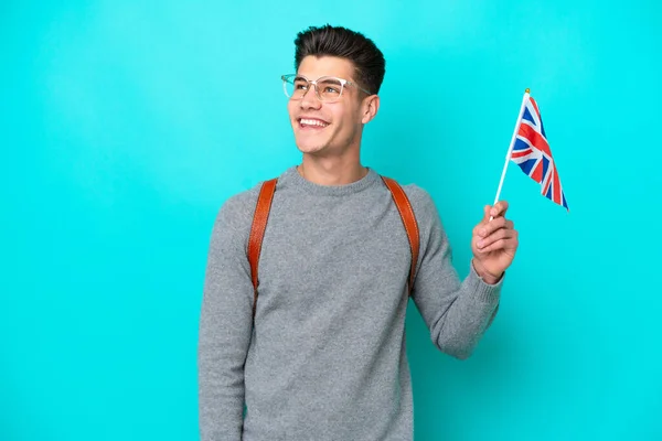 年轻的高加索人拿着一面英国国旗 蓝色背景隔离 一边高瞻远瞩 一边思考着一个想法 — 图库照片