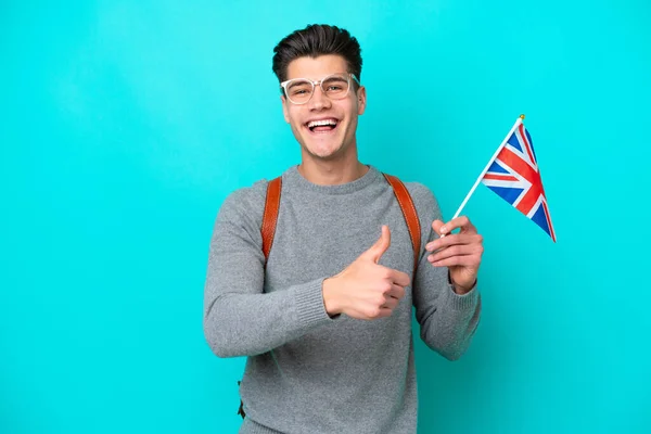 Young caucasian man holding an United Kingdom flag isolated on blue background giving a thumbs up gesture