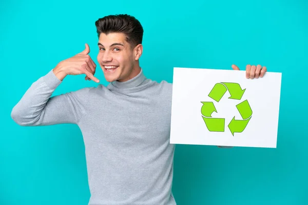 Young handsome caucasian man isolated on blue bakcground holding a placard with recycle icon and doing phone gesture