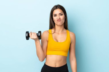 Beyaz bir kadın halter kaldırıyor mavi arka planda hüzünlü bir ifadeyle.