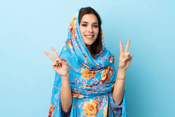 両方の手で勝利のサインを示す青の背景に隔離された伝統的な衣装を持つ若いモロッコの女性 — ストック写真