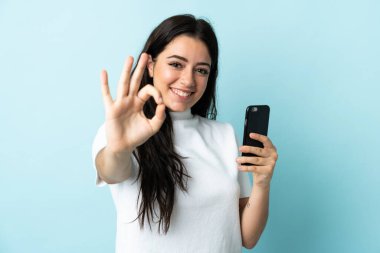 Mavi arkaplanda izole edilmiş cep telefonu kullanan genç bir kadın parmaklarıyla tamam işareti gösteriyor