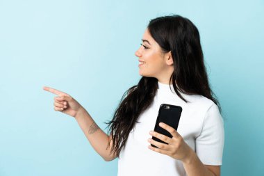 Mavi arka planda izole edilmiş cep telefonu kullanan genç bir kadın bir ürünü sunmak için kenarı işaret ediyor