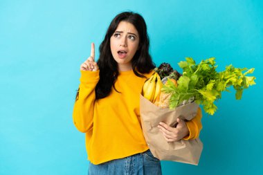 Elinde alışveriş torbası tutan genç bir kadın mavi arka planda izole edilmiş bir şekilde parmağı işaret eden bir fikir düşünüyor.