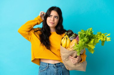 Elinde alışveriş çantası tutan genç bir kadın mavi arka planda izole edilmiş şüpheleri ve düşünceleri var.