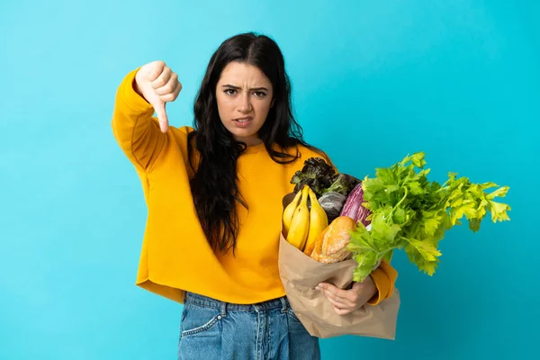 Elinde alışveriş poşeti tutan genç bir kadın mavi arka planda izole edilmiş, olumsuz bir ifadeyle baş parmağını gösteriyor.