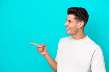 Genç, yakışıklı, beyaz bir adam, mavi bir bakkalda izole edilmiş bir şekilde, parmağını kenarı gösteriyor ve bir ürün sunuyor.