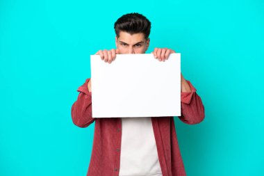 Genç, beyaz bir adam mavi bir bakkalda izole edilmiş boş bir levha tutuyor ve arkasında saklanıyor.