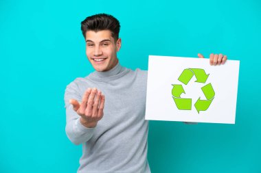 Mavi bakkalda izole edilmiş yakışıklı beyaz bir adam elinde geri dönüşüm simgesiyle bir pankart tutuyor ve bir jest yapıyor.