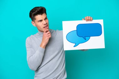 Genç, beyaz bir adam mavi bir bakkalda izole edilmiş. Elinde konuşma baloncuğu ikonu olan bir levha var ve düşünüyor.