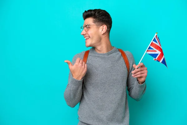 年轻的高加索男子举着一面英国国旗 蓝色背景隔离 指向侧面展示产品 — 图库照片