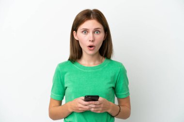 Genç İngiliz kadın beyaz arka planda izole edilmiş kameraya bakarken şaşırmış bir ifadeyle cep telefonunu kullanıyor.