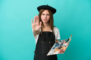 Mavi arka planda izole edilmiş palet tutan genç bir İngiliz kadın dur hareketi yapıyor.