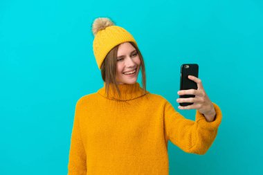Mavi arka planda izole edilmiş kışlık ceket giyen genç İngiliz kadın selfie çekiyor.