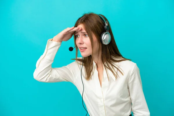 Telemarketer English woman working with a headset isolated on blue background looking far away with hand to look something