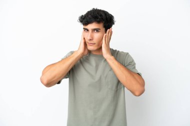 Beyaz arka planda izole edilmiş genç Arjantinli adam hayal kırıklığına uğramış ve kulaklarını kapatmış.