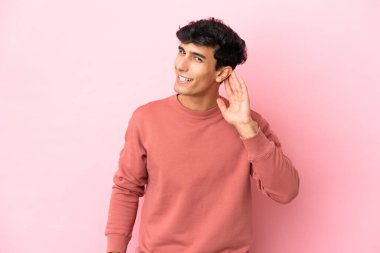 Pembe arka planda izole edilmiş genç Arjantinli bir adam bir şeyler dinlerken elini kulağına koyar.