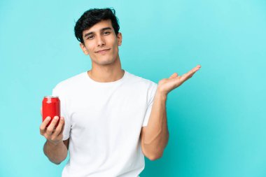 Arjantinli genç adam mavi arka planda izole edilmiş bir içecek tutuyor. Gelmesi için ellerini yana uzatıyor.