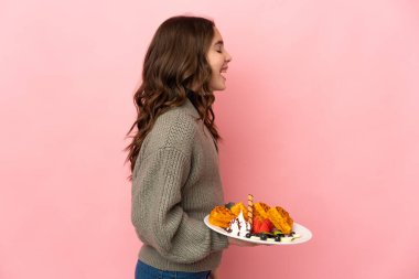 Pembe arka planda izole edilmiş waffle tutan küçük kız yan pozisyonda gülüyor.