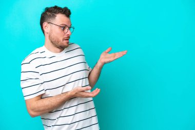 Genç Brezilyalı bir adam mavi arka planda izole edilmiş. Sürpriz bir ifadeyle yan tarafa bakıyor.