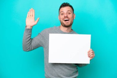 Elinde boş bir levha tutan genç Brezilyalı bir adam mavi arka planda izole edilmiş boş bir levha ve selam veriyor.