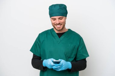 Yeşil üniformalı cerrah beyaz arka planda izole edilmiş cep telefonuyla bir mesaj gönderiyor.