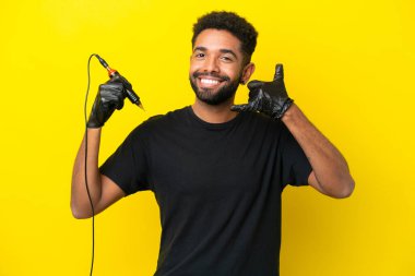 Dövme sanatçısı Brezilyalı adam sarı arka planda izole edilmiş, telefon jesti yapıyor. Bana geri dönüş işareti ver.