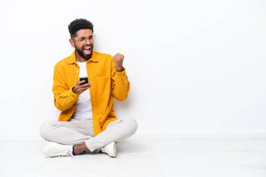 Yerde oturan Brezilyalı genç adam beyaz arka planda izole edilmiş ve telefon zafer pozisyonunda.