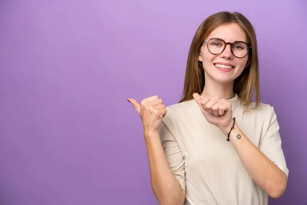 製品を提示するために側面を指す紫色の背景に隔離された若い英語の女性 — ストック写真