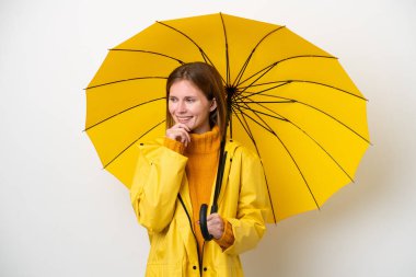 Yağmura dayanıklı ceketi ve şemsiyesi olan genç bir İngiliz kadın beyaz arka planda izole edilmiş, yana bakıyor ve gülümsüyor.