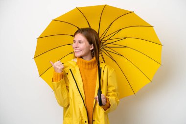 Yağmura dayanıklı ceketli ve şemsiyeli genç bir İngiliz kadın beyaz arka planda izole edilmiş bir ürün sunmak için yan tarafı işaret ediyor.