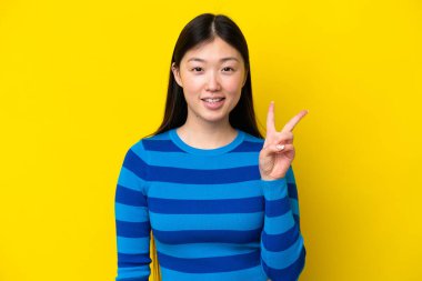 Sarı arka planda izole edilmiş genç Çinli kadın gülümsüyor ve zafer işareti gösteriyor.