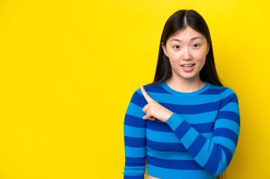 Sarı arka planda izole edilmiş genç Çinli kadın şaşırmış ve işaret ediyor.