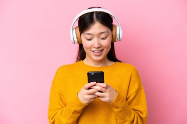 Genç Çinli bir kadın pembe arka planda izole edilmiş müzik dinliyor ve cep telefonu arıyor.